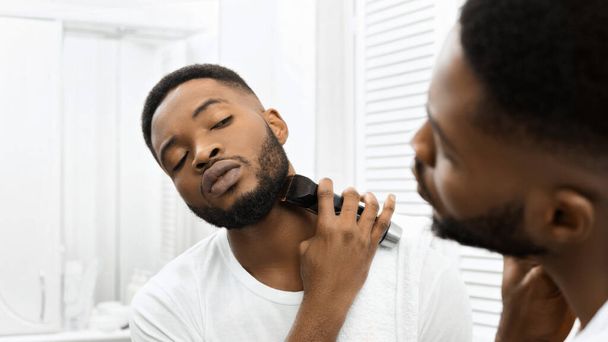 Фокусований афроамериканський хлопець тримає бороду, використовуючи тримери в добре освітленій, сучасній обстановці ванної кімнати - Фото, зображення