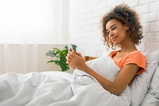 Una joven afroamericana yace cómodamente en la cama, comprometida con su smartphone, retratando una sensación de ocio y conexión digital - Foto, imagen