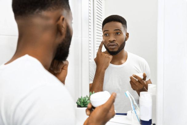 Africký Američan je vidět, jak nanáší krém na obličej, zaměřuje se na péči o jeho kůži při pohledu do zrcadla - Fotografie, Obrázek