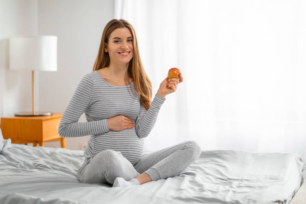 Une femme enceinte joyeuse s'assoit les jambes croisées sur un lit tenant une pomme, favorisant de saines habitudes alimentaires pendant la grossesse - Photo, image