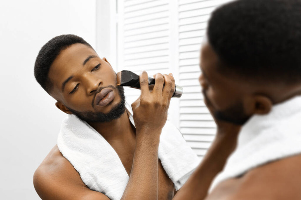 Afro-Américain avec une serviette autour du cou toilette sa barbe à l'aide d'une tondeuse électrique devant un miroir de salle de bain, axée sur l'autosoin - Photo, image
