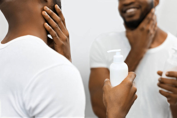 Africký Američan muž postava zkoumá produkt péče o pleť v koupelně, odraz ukazuje spokojenost s produktem - Fotografie, Obrázek