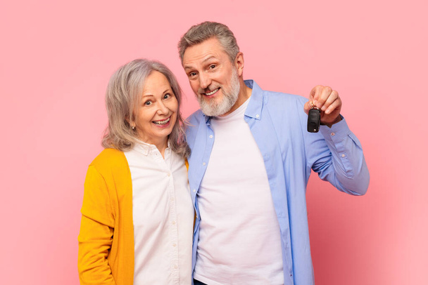 Šťastní důchodci pár nadšeně předvádět své nové auto klíč na růžovém pozadí, zralý manžel a manželka kupující rodinné auto společně v jejich odchodu do důchodu pro mobilitu a svobodu - Fotografie, Obrázek