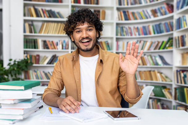 Cheerful young man with curly hair, waving and talking during a video call, surrounded by books at home. - Foto, afbeelding