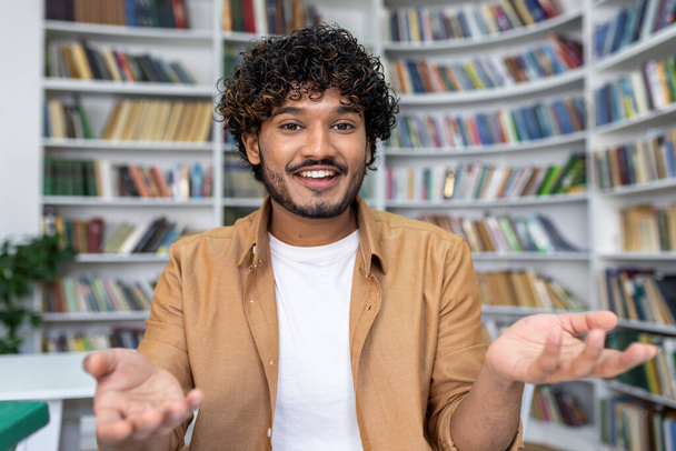 Cheerful young man with curly hair engaged in a video call, looking at camera, with a bookshelf background. - Foto, Imagem