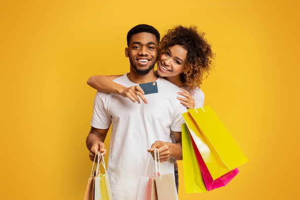 Cheerful african-american couple of shopaholics satisfied with their bank, embracing with credit card and shopping bags on orange background - Photo, Image