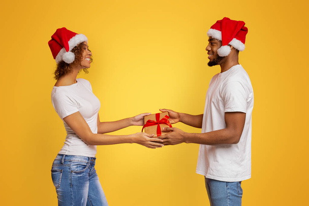 Familie cadeautjes uitwisselen. zwart millennial paar geven geschenk doos naar elkaar, oranje studio achtergrond - Foto, afbeelding