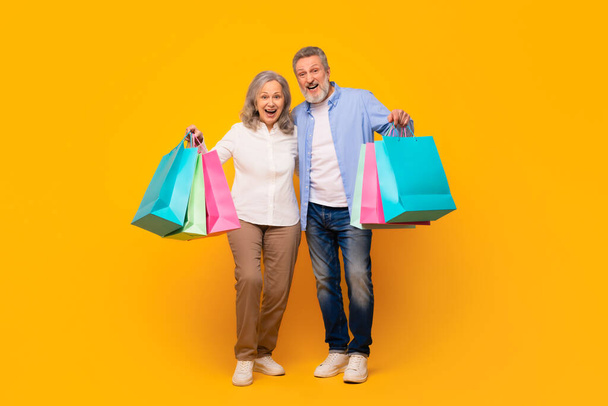 Happy senior husband and wife carrying colorful shopping bags, celebrating Black Friday sales, standing together with purchases against yellow studio backdrop. Mature consumerism. Full length - Photo, Image