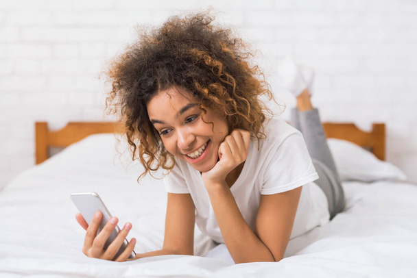 Genç kıvırcık saçlı Afro-Amerikalı bir kadın derin derin bir ifadeyle yatakta kendini desteklerken dikkatle cep telefonuna bakıyor. - Fotoğraf, Görsel