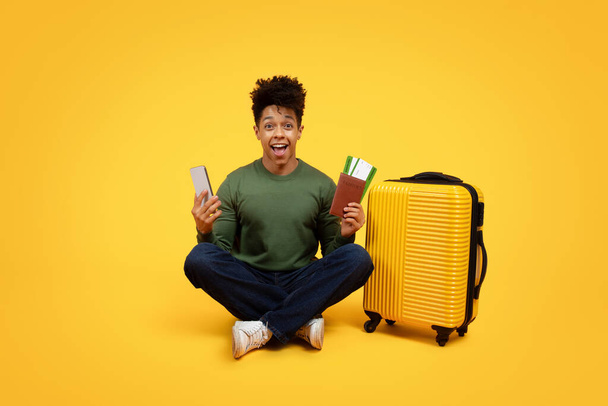 Un giovane afroamericano siede sul pavimento con il telefono in una mano e il passaporto con carte d'imbarco nell'altra, accanto a una valigia gialla brillante, esprimendo eccitazione, gioia per il viaggio - Foto, immagini