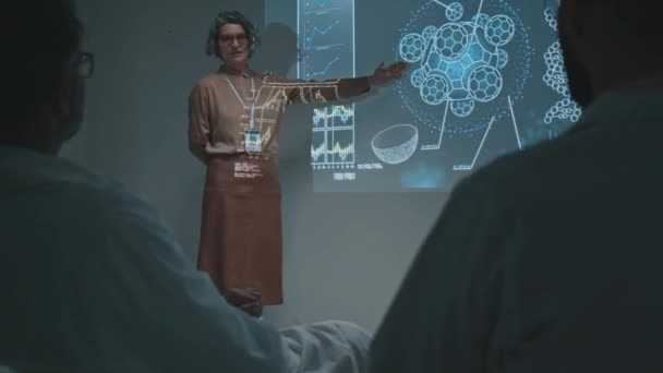 Середній повноцінний знімок кавказького лікаря середнього віку, що стоїть поруч з екраном проектора на засіданні кафедри, розповідає колегам про нову терапію, її дію та ефект - Кадри, відео