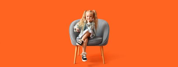 klein meisje in fauteuil kijken tv op oranje achtergrond - Foto, afbeelding