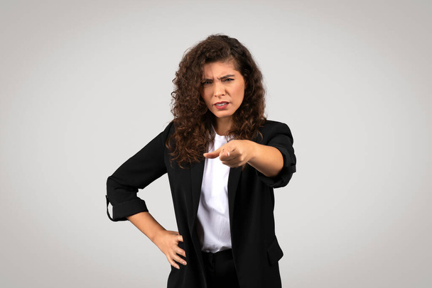 Eine Frau im Business-Anzug zeigt mit zweifelndem oder fragendem Gesichtsausdruck direkt in die Kamera, vor einem neutralen Hintergrund - Foto, Bild