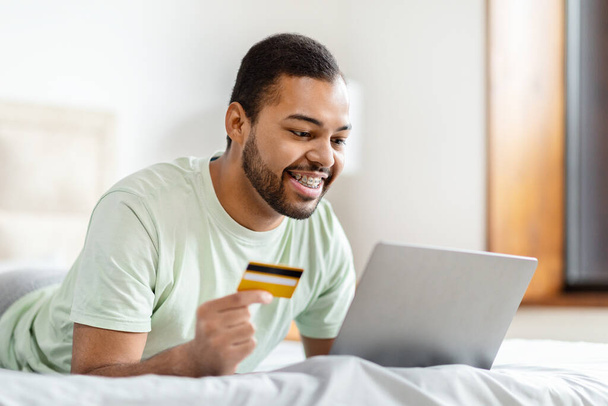 Junger afrikanisch-amerikanischer Mann mit Kreditkarte, Laptop, Online-Shopping oder Bezahlkonzept im häuslichen Umfeld - Foto, Bild