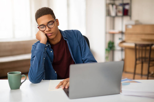 Vervelende tiener het dragen van een bril toont verveling tijdens het gebruik van een laptop op een kantoor thuis met een kopje koffie - Foto, afbeelding
