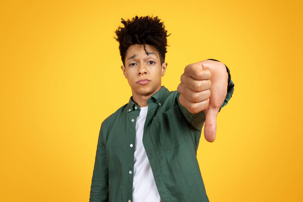 Ontevreden jonge Afro-Amerikaanse man toont duimen naar beneden gebaar met een sceptische blik op een gele achtergrond - Foto, afbeelding