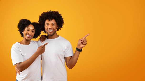 Milující tisíciletý africký americký pár v bílých tričkách objímající a ukazující ukazující prázdné kopírovací prostor pro reklamu, žluté pozadí studia, panorama - Fotografie, Obrázek