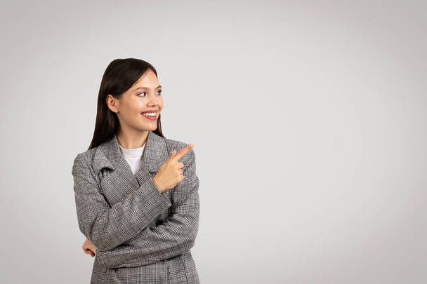 Fröhliche Geschäftsfrau im klassischen Tweed-Blazer, die mit zahmem Lächeln zur Seite zeigt und vor schlichtem grauen Hintergrund Wahl oder Richtung andeutet - Foto, Bild