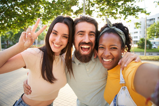 Selfie tři nadšené multiraciální radostné mladé přátele objímající venku. Ženy a muž se baví při pohledu na usmívající se fotoaparát a dělat mír symbol ruce těší letní dovolenou. Gen z ve slunečném parku - Fotografie, Obrázek