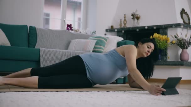 Terhes anya a harmadik trimeszterben részt vesz otthon Terhességi edzés, fekvő padló matrac a nappaliban, követő gyakorlatok tabletta eszköz - Felvétel, videó