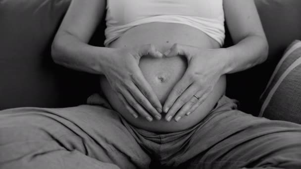 Amor sincera monocromática de la madre haciendo símbolo del corazón en la parte superior del vientre embarazada durante la última etapa del embarazo en blanco y negro - Imágenes, Vídeo