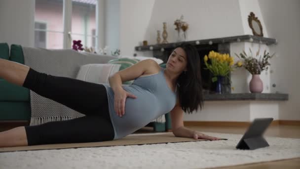 Hamile bir kadın hamileliğinin üçüncü üç aylık döneminde masa başında dijital koçu takiben evdeki oturma odasının zeminine uzanmış doğum öncesi egzersizleri yapıyor. - Video, Çekim