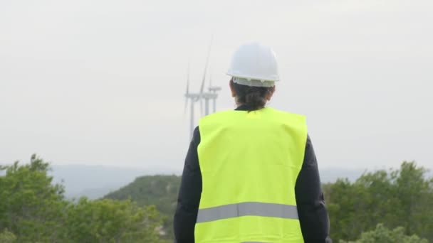 Vista trasera de la ingeniera de electricidad femenina con uniforme y casco mirando turbinas eólicas en las montañas - Imágenes, Vídeo