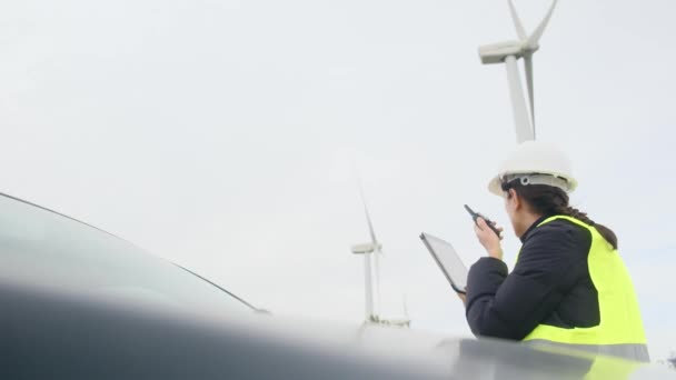 Ingegnere elettrico femminile vicino auto parla su walkie-talkie, utilizza tablet digitale e controllo turbine eoliche - Filmati, video