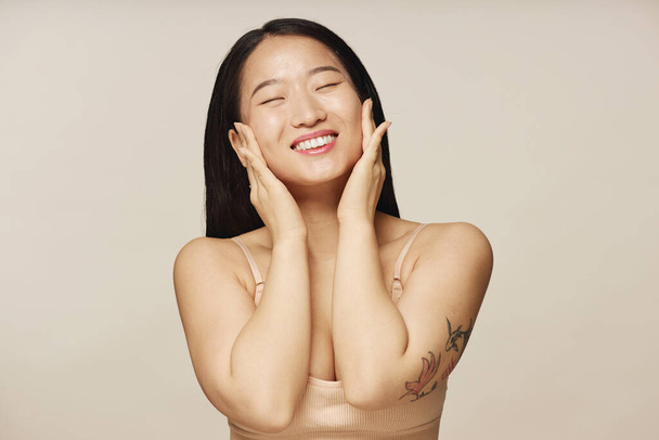 Chest up studio portrait of happy young Asian woman wearing beige bra enjoying touching her smooth facial skin - Foto, immagini