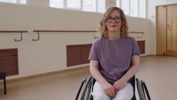 Портрет веселої блондинки сидить у інвалідному візку і позує на камеру з посмішкою в танцювальній студії - Кадри, відео
