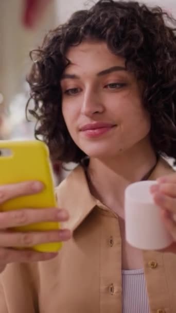 Vertikale Nahaufnahme einer jungen kaukasischen Frau mit lockigem kaukasischem Haar, die ein Glas Creme in der Hand hält und per Smartphone nach Bewertungen im Internet sucht, während sie sich für eine Hautpflege im Geschäft entscheidet - Filmmaterial, Video