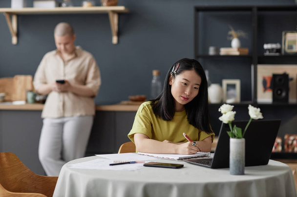 若いアジアの大学生は,オンライン講義を見て自宅でダイニングテーブルに座ってメモを作り,彼女のガールフレンドは背景に立っています - 写真・画像