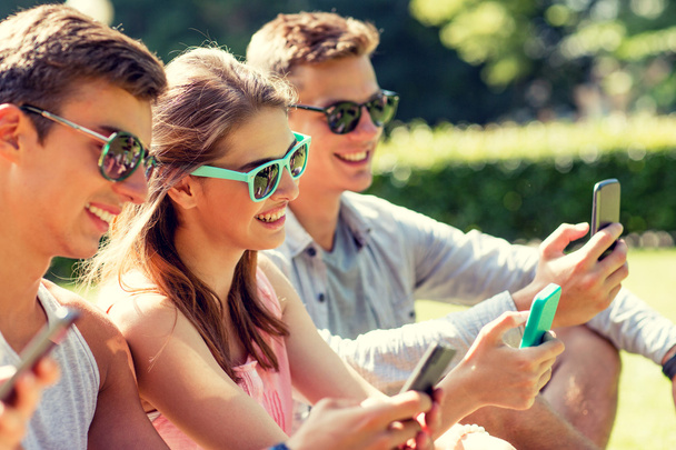 smiling friends with smartphones sitting on grass - 写真・画像