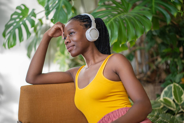 Rahat Afro-Amerikan kadın ev bahçesinde dinleniyor kablosuz kulaklık kullanıyor müzik dinliyor hafta sonları evcil bitkilerle çevrili. Yeşil odada stresi azaltmak için siyah genç bir kadının dinlendiğini hayal etmek. - Fotoğraf, Görsel