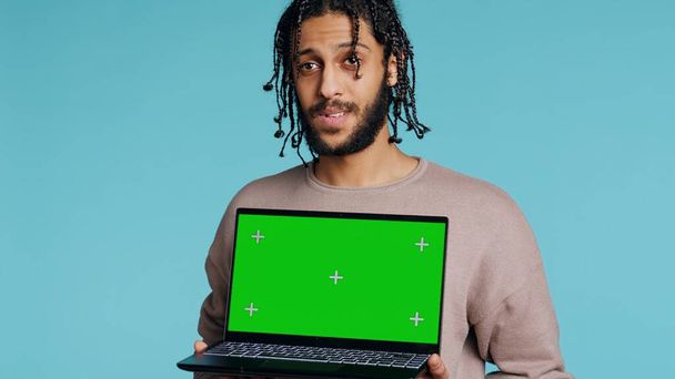 Homem BIPOC alegre segurando laptop tela verde, tendo humor positivo. Pessoa sorridente apresentando croma key notebook, fazendo recomendação, isolado sobre o fundo do estúdio, câmera B - Foto, Imagem