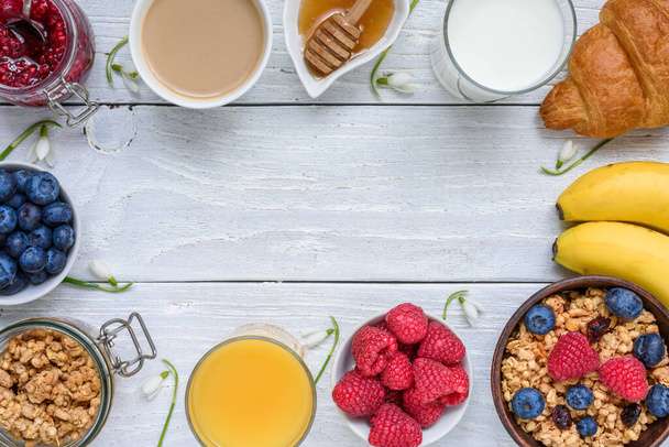 Здоровий сніданок "шведський стіл" з різними ранковими стравами та весняними квітами. Рамка з граноли, кави, фруктів, ягід, молока, апельсинового соку та меду на білому дерев'яному столі. Вид зверху з пробілом для копіювання. - Фото, зображення