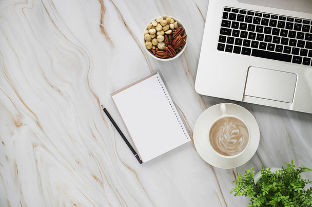 Top view photo of workplace with laptop pencil, paper notebook, plant, cup of coffee and mixed nuts in bowl on white marble table background. - Photo, Image