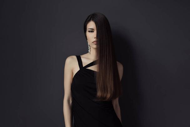Ohromující portrét stylové ženy s dlouhými rovnými vlasy v elegantních černých šatech a elegantních náušnicích, pózující na tmavém pozadí. - Fotografie, Obrázek