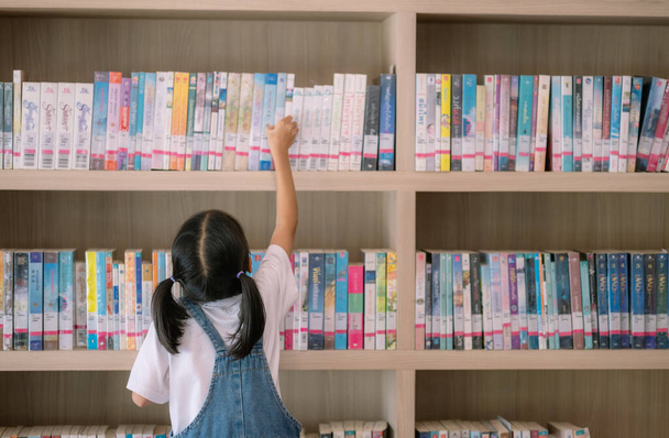 Młoda dziewczyna sięga po książkę na półce bibliotecznej. Biblioteka jest wypełniona wieloma książkami, a dziewczyna skupia się na znalezieniu tej, której chce. Pojęcie ciekawości i uczenia się - Zdjęcie, obraz