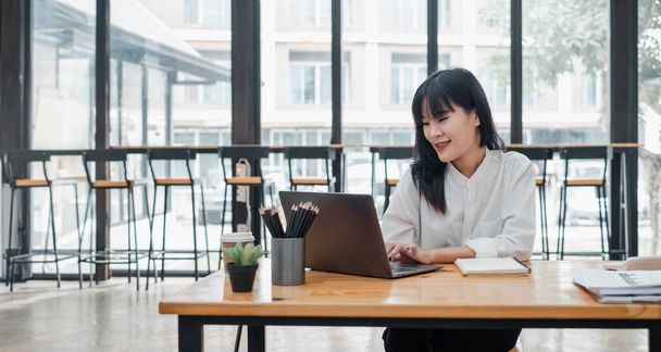 Mujer asiática sonriente centrada en su trabajo portátil en un ambiente de cafetería brillante con café y papelería en la mesa. - Foto, imagen