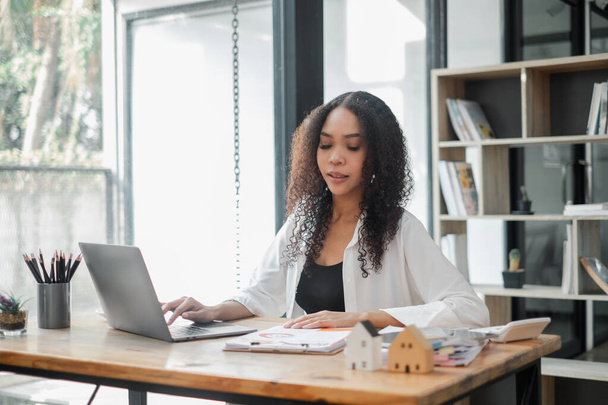 Professionele vrouw gericht op haar laptop, omgeven door georganiseerde werkdocumenten en modellen van huizen op haar bureau in een helder kantoor. - Foto, afbeelding