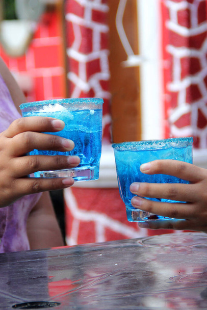 Δύο νεαρές μελαχρινές Λατίνες γυναίκες 20 ετών πίνουν παγωμένο μπλε κοκτέιλ με τσίλι, γιορτάζουν πίνοντας αλκοόλ - Φωτογραφία, εικόνα