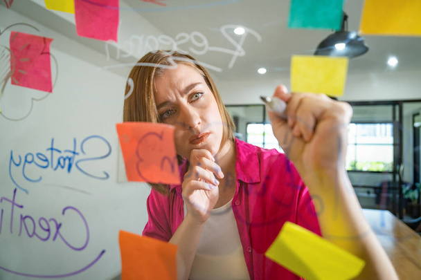 Closeup of young beautiful caucasian business leader presents marketing idea while writing marketing idea on glass board with mind map and colorful sticky notes. Portrait. Brainstorm. Immaculate. - Photo, Image