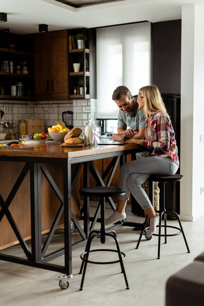 Веселая пара наслаждается беззаботным моментом на своей солнечной кухне, работая на ноутбуке в окружении здорового завтрака - Фото, изображение