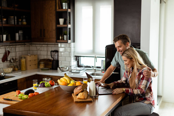 Una coppia allegra gode di un momento spensierato nella loro cucina soleggiata, lavorando su un computer portatile circondato da una colazione sana - Foto, immagini