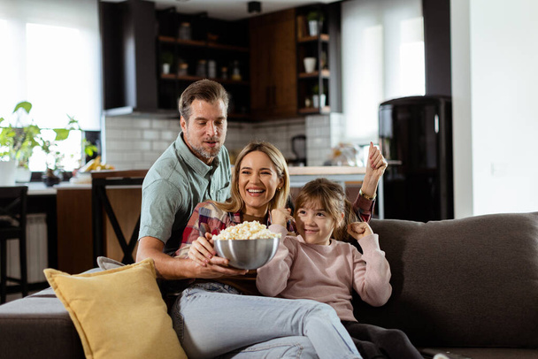Eine dreiköpfige Familie liegt gemütlich auf einer Couch, ihre Gesichter spiegeln Aufregung und Aufmerksamkeit wider, während sie sich während eines spannenden Filmabends eine Schüssel Popcorn teilen. - Foto, Bild