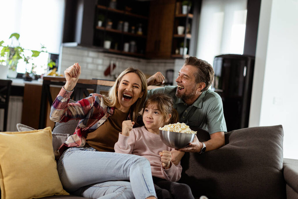 Una famiglia di tre persone è comodamente adagiata su un divano, i loro volti riflettono eccitazione e attenzione mentre condividono una ciotola di popcorn durante una serata cinematografica sospesa. - Foto, immagini