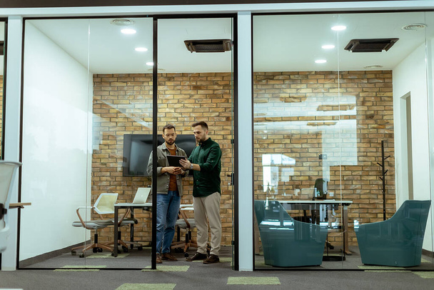 Два профессионала беседуют, стоя в офисном зале, в окружении элементов современного дизайна - Фото, изображение