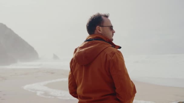 Pohled zezadu na muže ve slunečních brýlích stojícího na pobřeží oceánu a ráno sledujícího vlnící se vodu. Mužská procházka po mořském pobřeží venku a užívat si samoty během dovolené nebo víkendu - Záběry, video