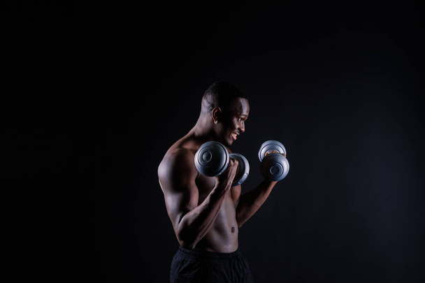 Isolated african muscular man with dumbbells on a dark studio background. Strong shirtless black guy - Foto, Imagem